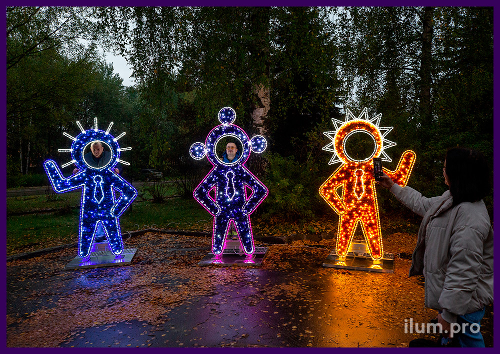 Три светящихся фигуры с отверстиями для лица из гирлянд и мишуры в парке